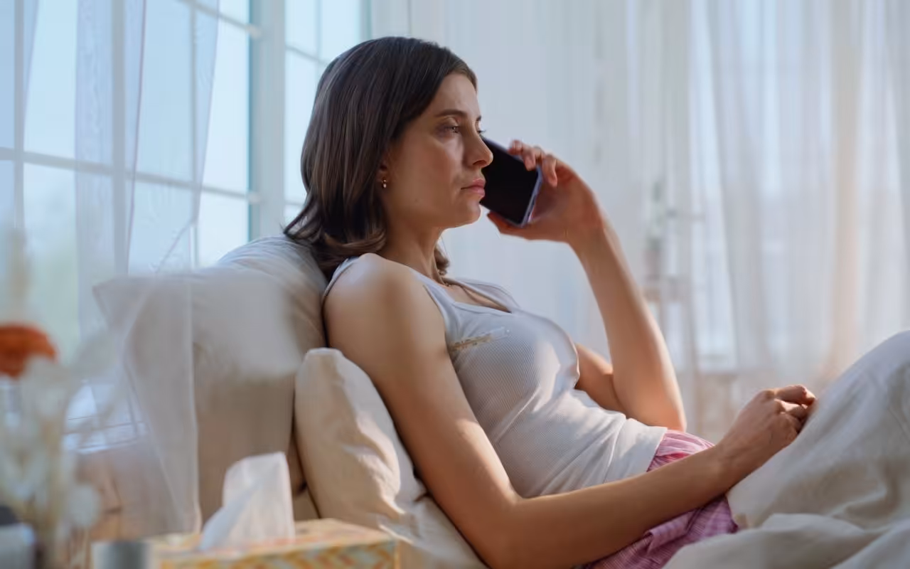 A sick woman in bed holds a phone to her ear with a thermometer under her arm, calling off work on short notice.