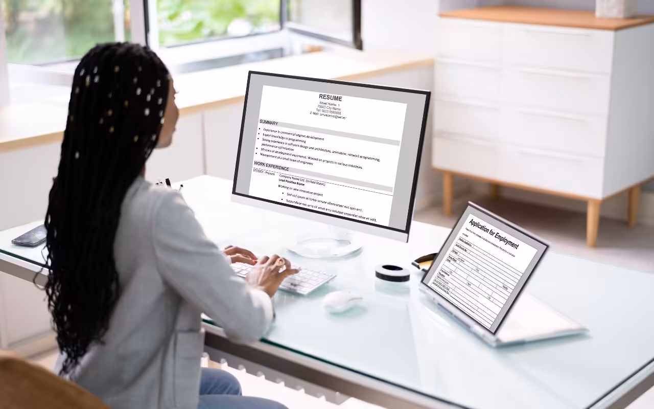 A person types a resume on a large desktop monitor while a job application is open on a nearby laptop.